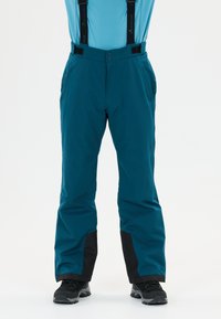 Türkise Schneehose mit schwarz verstärkten Säumen, verstellbaren Trägern und schwarzen Wanderschuhen an einer Person, die vor einem weißen Hintergrund steht.
