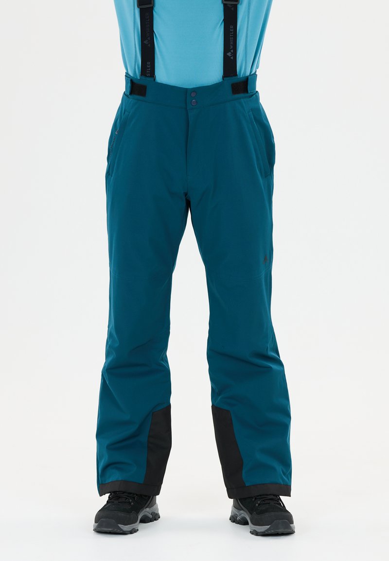 Türkise Schneehose mit schwarz verstärkten Säumen, verstellbaren Trägern und schwarzen Wanderschuhen an einer Person, die vor einem weißen Hintergrund steht.