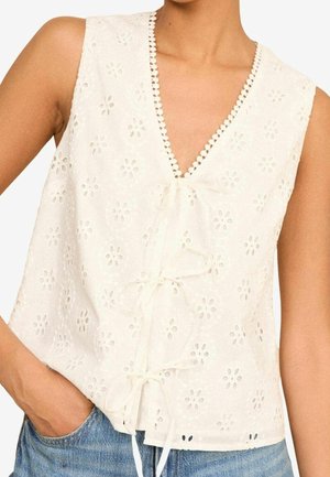 Biała bezrękawna bluzka eyelet z kwiatowymi wycięciami, dekoltem w kształcie litery V z falistą lamówką i trzema wiązaniami z przodu, noszona z niebieskimi dżinsami.