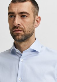 Rövid, sötét hajú és szakállas férfi, aki világoskék gombos inget visel, sima világos háttér előtt.