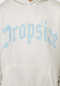 Hvit hettegenser med lyseblå gotisk tekst "Dropsize" på forsiden. Materialet ser mykt ut og har en lommesekk foran samt hette med snøring.