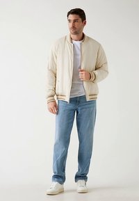 Veste bomber matelassée beige clair avec poignets et ourlet côtelés, portée sur un t-shirt blanc, associée à un jean bleu et des baskets blanches.