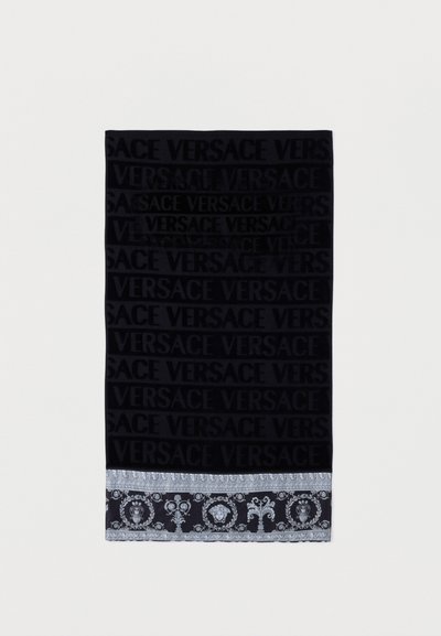 Μαύρη πετσέτα με επαναλαμβανόμενο κείμενο "VERSACE" σε βελούδο και διακοσμητικό ασημένιο μπαρόκ μοτίβο στο τελείωμα.