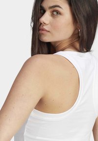 Débardeur blanc sans manches avec un col rond et une coupe ajustée, mettant en valeur une texture en coton lisse et une silhouette au dos ouvert.