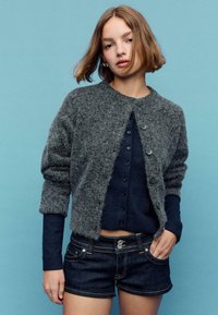 Sivi, puhasti cardigan z okroglim izrezom in gumbi spredaj, oblečen čez mornarsko modro zgornjo majico z gumbi, kombiniran s temnimi denim kratkimi hlačami.