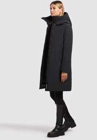 Sort lang frakk med hætte, med lynlås og trykknapslukning, sidelommer, glat tekstur, parret med sorte støvler, der går op til knæet.