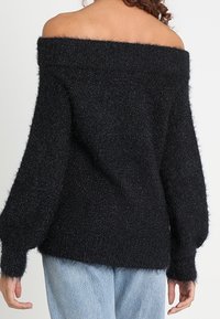 Czarny sweter z odkrytymi ramionami o puszystej teksturze, szerokich rękawach i luźnym kroju. Posiada prążkowany dekolt i miękki, gruby materiał.
