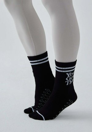 Nogi w czarnych skarpetach antypoślizgowych z białymi paskami i napisem „You Got This”, stojące na palcach na jednolitym jasnym tle.