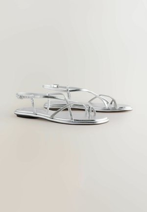 Srebrne ravne sandale s metalik trakama, koje imaju ukrštene dizajne i podesive gležnjeve. Glatka tekstura i dizajn otvorenih prstiju.