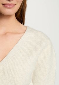 Maglione lavorato a maglia di colore crema con scollo a V, tessuto testurizzato e leggero lucido, caratterizzato da una vestibilità ampia e dettagli a costine sui bordi.