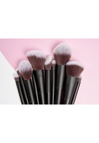Set von Makeup-Pinseln mit schwarzen Griffen und weichen Borsten, die an den Spitzen einen Farbverlauf von Dunkelbraun zu Hellrosa aufweisen, auf einem rosa Hintergrund angeordnet.