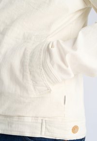 Jasnobeżowa kurtka wykonana z miękkiej tkaniny, z boczną kieszenią, detalem z guzików i widocznymi wzorami przeszyć. Luźny fason z podwiniętymi rękawami.