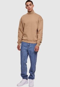 Felpa beige a collo alto con spalle scese, abbinata a jeans azzurri chiari con vestibilità comoda e tasche applicate. Indossata con sneakers bianche.