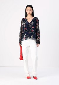 Blouse florale marine avec col froncé, manches transparentes ; associée à un pantalon blanc, un sac à main rouge et des escarpins rouges à bout pointu, sur un fond clair.
