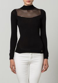 Frau in einem enganliegenden schwarzen Langarmshirt mit einem Netzpaneel über der Brust, kombiniert mit weißen Hosen vor einem schlichten Hintergrund.