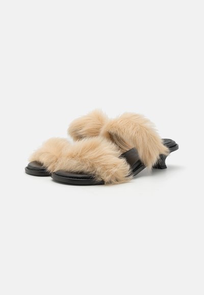 Faux fur slides with beige fluffy material and black leather base. Features dual fur straps and a curved heel for added style.