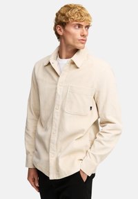 Giacca camicia in corduroy beige con chiusura a bottoni, taschino sul petto e colletto. Presenta righe verticali e maniche lunghe.
