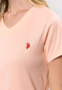 Hell pfirsichfarbenes Baumwoll-T-Shirt mit V-Ausschnitt, kurzen Ärmeln und einem kleinen roten, gestickten Logo auf der linken Brust. Weiche Textur und lässige Passform.