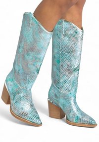 Botas de serpiente en azul y plata con punta afilada, altura hasta la mitad de la pantorrilla y un tacón de bloque de madera. Costuras blancas que realzan el diseño.