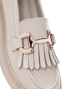 Mocassin en cuir beige clair avec une boucle décorative en ton doré et un détail en frange. Présente une couture contrastante et une semelle texturée.