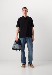 Sort polo-shirt med korte ærmer, holdt med en denimjakke i hånden; parret med lyseblå jeans og brune sko, stående mod en ensfarvet baggrund.
