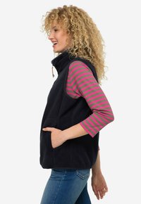 Granatowa kamizelka polarowa z przednimi kieszeniami, z wysokim kołnierzem oraz koszulą z długim rękawem w paski w kolorze oliwkowym i różowym. Widoczne kręcone włosy.