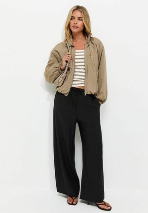 Jonge vrouw met blond haar die een beige jas, een gestreepte crop top, wijde zwarte broek en zwarte sandalen met hakken draagt, en een beige handtas vasthoudt.