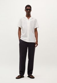 Camisa blanca de manga corta con rayas verticales, botones amarillos, combinada con pantalones holgados gris oscuro y sandalias negras.