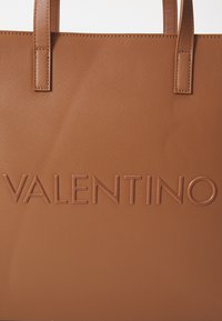 Brun texturerad toteväska med dubbla handtag och upphöjd "VALENTINO"-logotyp präglad över framsidan.