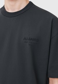 Sort bomulds T-shirt med korte ærmer, rund halsudskæring og præget 'ALLSAINTS' logo på venstre brystområde. Glat tekstur.