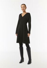 Schwarzes langärmliges Kleid mit V-Ausschnitt und schwingendem Rockdesign, aus einem strukturierten Stoff gefertigt. Getragen mit enganliegenden schwarzen Strumpfhosen und kniehohen Stiefeln.