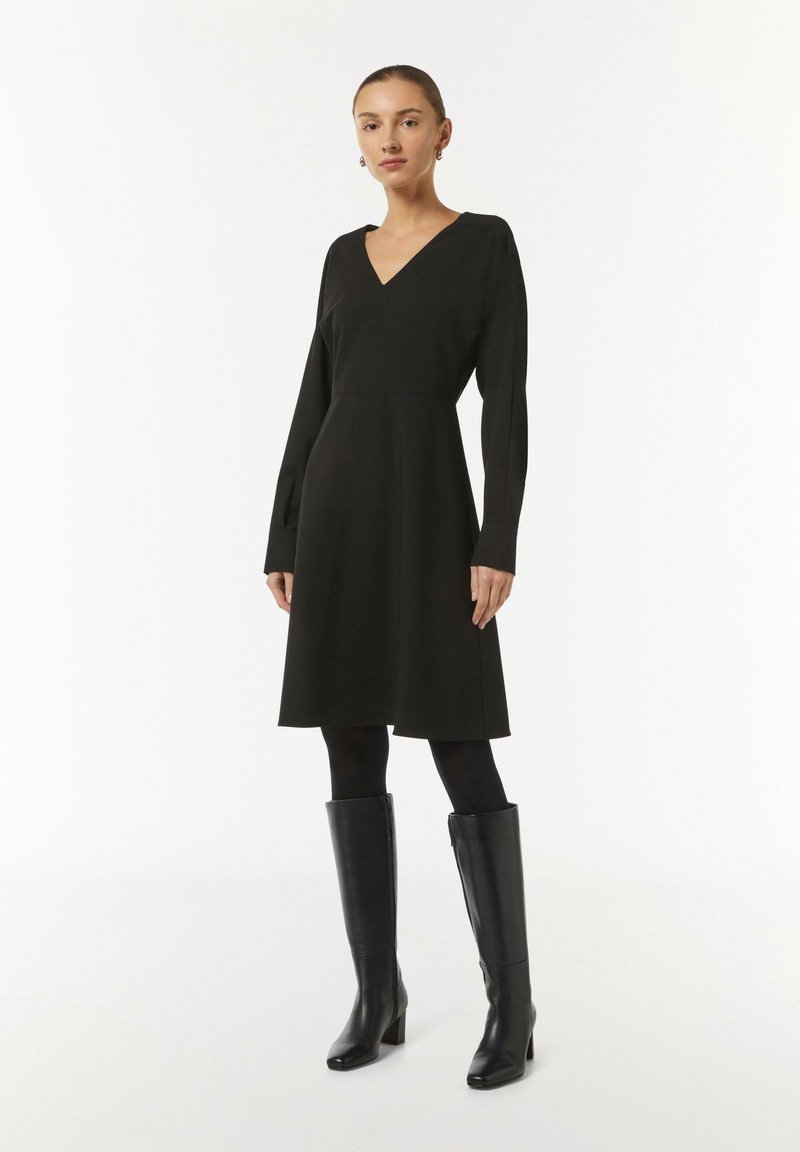 Schwarzes langärmliges Kleid mit V-Ausschnitt und schwingendem Rockdesign, aus einem strukturierten Stoff gefertigt. Getragen mit enganliegenden schwarzen Strumpfhosen und kniehohen Stiefeln.