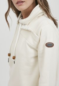 Crème-kleurige hoodie met een bloemenpatroon aan de binnenkant, voorzien van een hoge kraag, verstelbare koordjes met houten toggles en een logopatch.