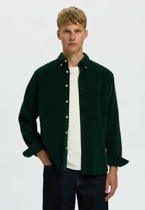 Homme blond portant une chemise boutonnée vert foncé sur un T-shirt blanc et un jean foncé, debout avec une main dans la poche devant un fond uni.