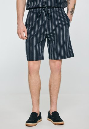 Mężczyzna w czarno-białych szortach w paski ze sznurkiem, czarne buty wsuwane, stoi z rękami w kieszeniach na jednolitym tle.