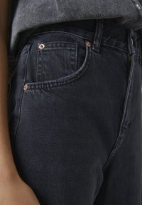 Lähikuva tummansinisten farkkujen etupussista, näkyvistä kuparin niiteistä, vyölenkeistä ja sisätaskussa olevasta harmaasta t-paidasta.