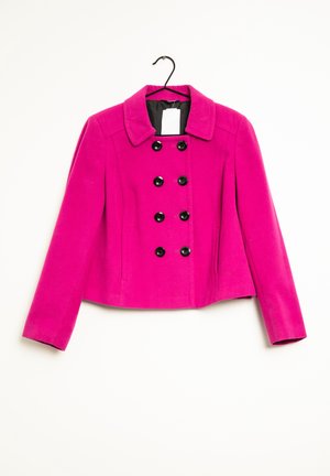 Lyserødt, dobbeltspændt kvindekåbe med sorte knapper hængende på en sort bøjle mod en hvid baggrund.