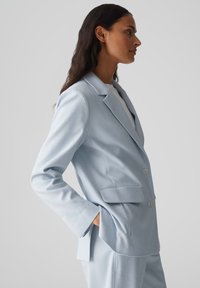 Blazer su misura di un azzurro chiaro con revers a punta, due bottoni e tasche frontali. Realizzato in un tessuto liscio con un design strutturato.
