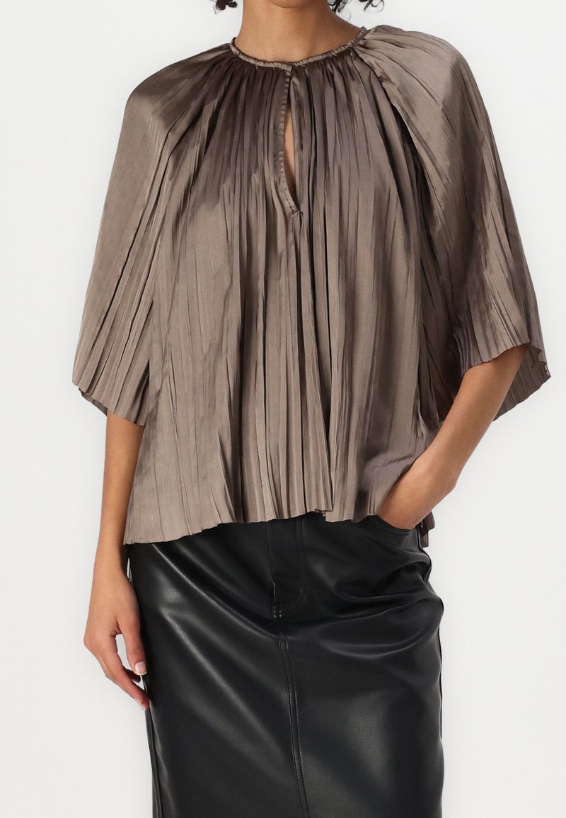 Femme portant un haut plissé taupe avec un décolleté en trou de serrure et des manches mi-longues, associé à une jupe crayon en cuir noir.