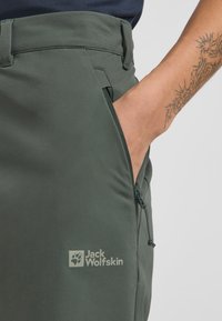 Dunkelgrüne Cargo-Hosen mit glatter Textur, ausgestattet mit einer Reißverschlusstasche und einem sichtbaren Logo-Druck an der Seite.