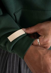 Manos ajustando la manga de una sudadera verde oscura con un parche rectangular beige y un anillo plateado en la mano derecha.