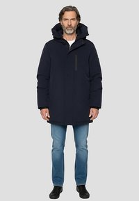 Donkerblauwe gewatteerde jas met capuchon, voorzien van een voorrits, zijzakken en een iets langere snit, gedragen over lichtblauwe spijkerbroek en zwarte schoenen.