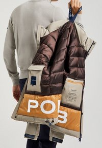 Osoba držící béžovou a hnědou prošívanou bundu s velkým textem "POLO" na hořčicovém panelu a viditelnými několika vnitřními kapsami.