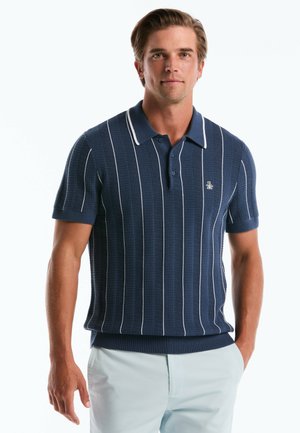 Mann in einem marineblauen, kurzärmeligen, gestrickten Poloshirt mit weißen vertikalen Streifen und hellen Hosen, steht mit einer Hand in der Tasche.