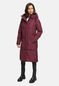 Woman wearing long burgundy quilted winter coat with hood, black leggings, and black mid-calf boots, standing with one hand in pocket.