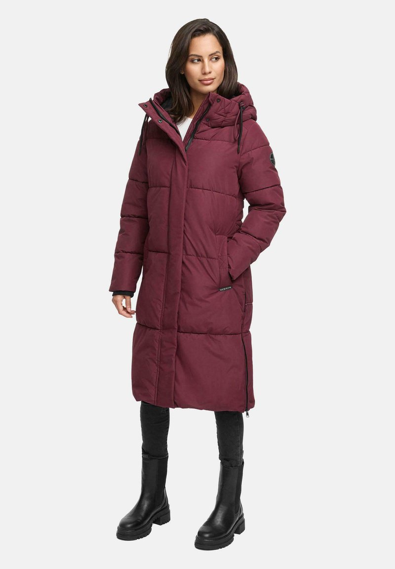 Woman wearing long burgundy quilted winter coat with hood, black leggings, and black mid-calf boots, standing with one hand in pocket.