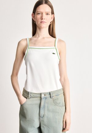 Witte tanktop met vierkante halslijn, dunne bandjes en groene afwerking. Beschikt over een klein logo aan de linkerzijde. Gepaard met lichtblauwe denim broek.
