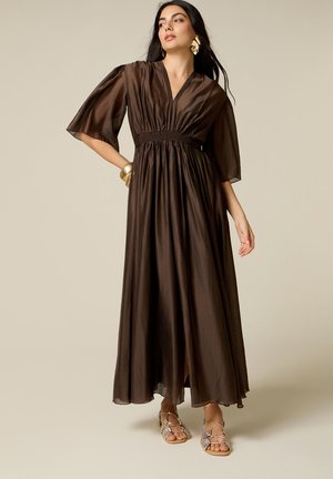 Frau trägt ein fließendes dunkelbraunes Maxikleid mit V-Ausschnitt, halblangen Puffärmeln, goldenen Ohrringen und Armband sowie flache Sandalen aus Schlangenleder.