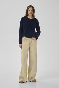 Frau steht vor grauem Hintergrund, trägt einen marineblauen Kapuzenpullover und weite beige Jeans mit beigen Schuhen.