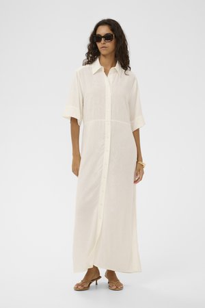 Woman wearing a long white button-down dress, dark sunglasses, tan heeled sandals, and gold bracelets stands against a plain background.